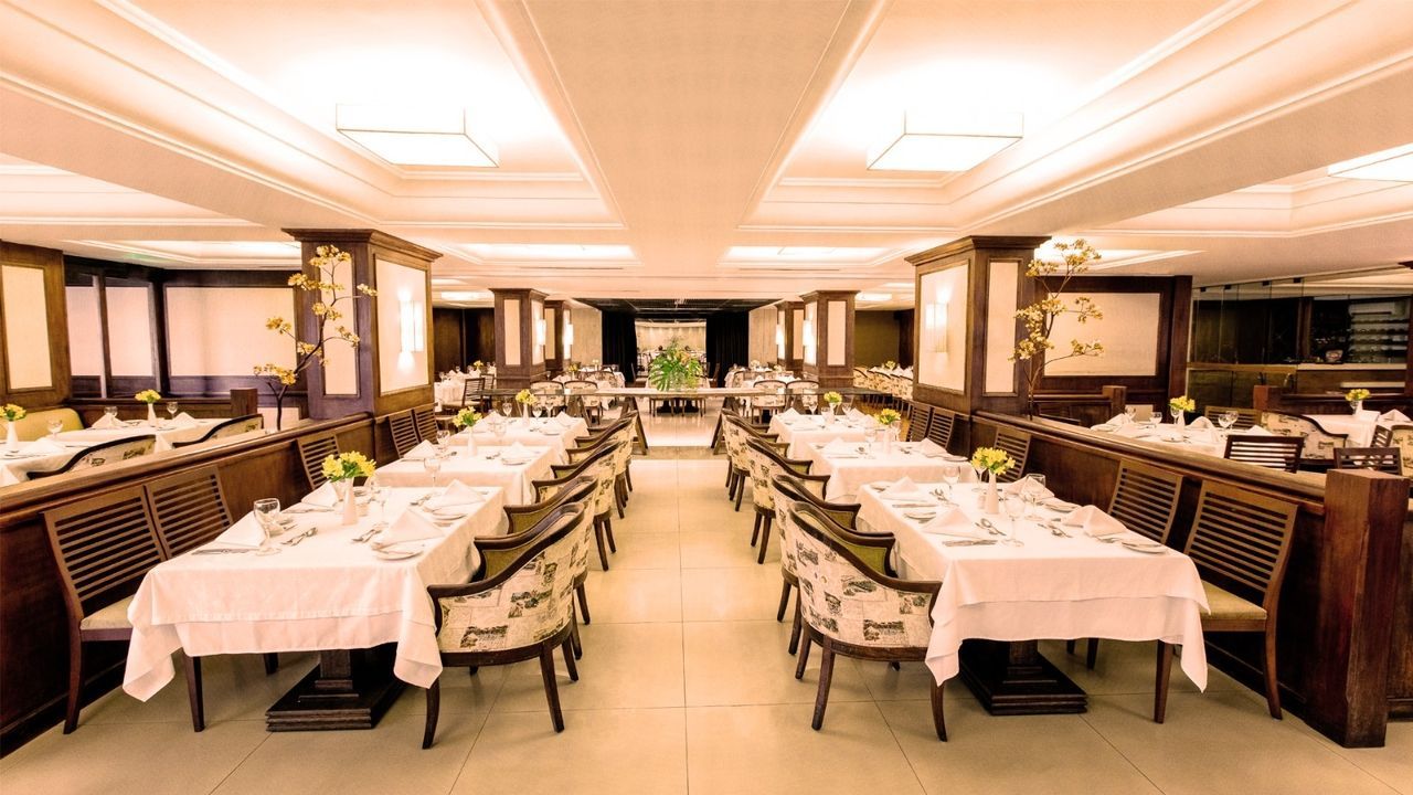 Sala de jantar formal com mesas compridas preparadas para refeições. Toalhas de mesa brancas, cadeiras estampadas e painéis de madeira.