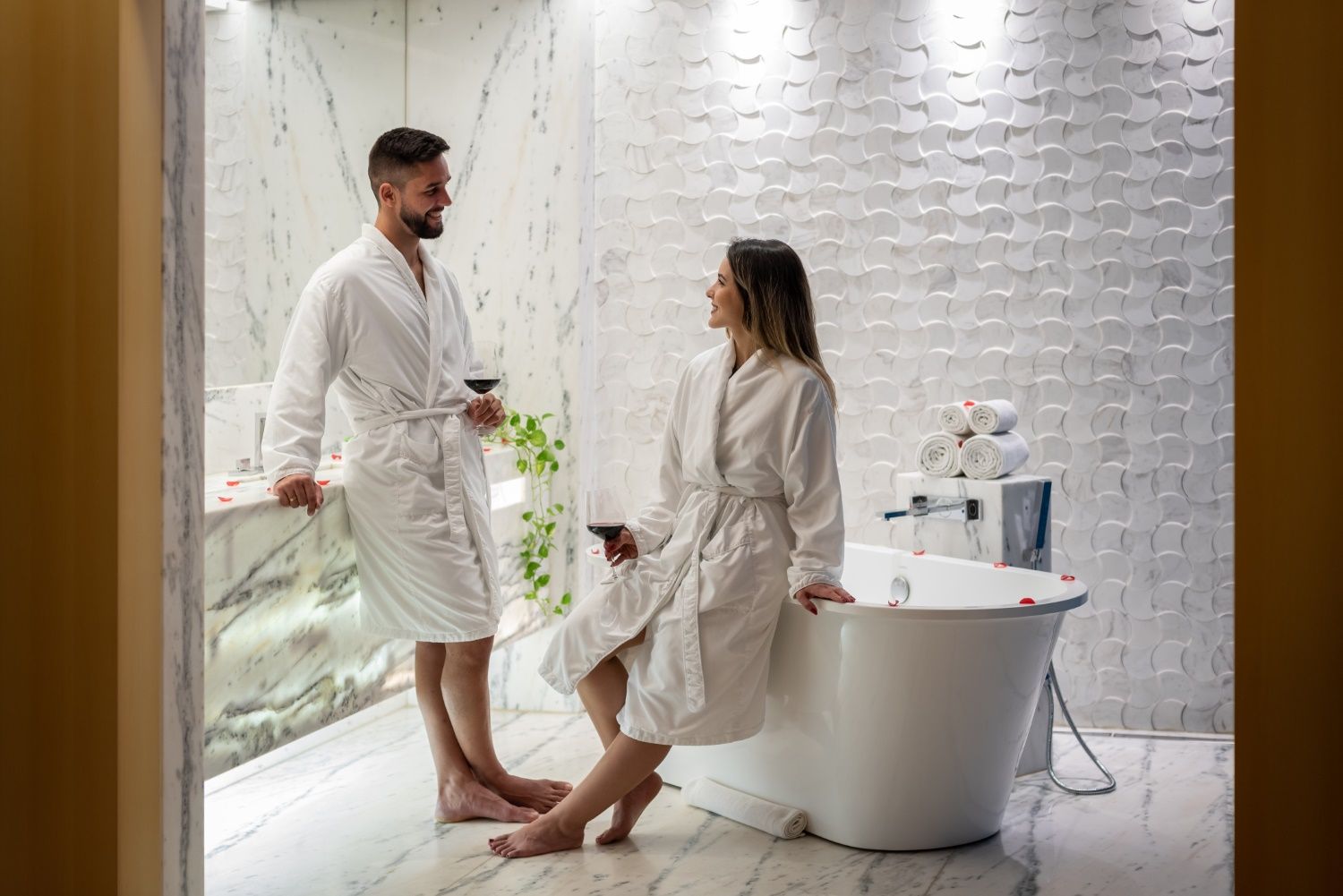 Um casal de robes, bebendo vinho, conversando perto de uma banheira em um banheiro estilo spa.