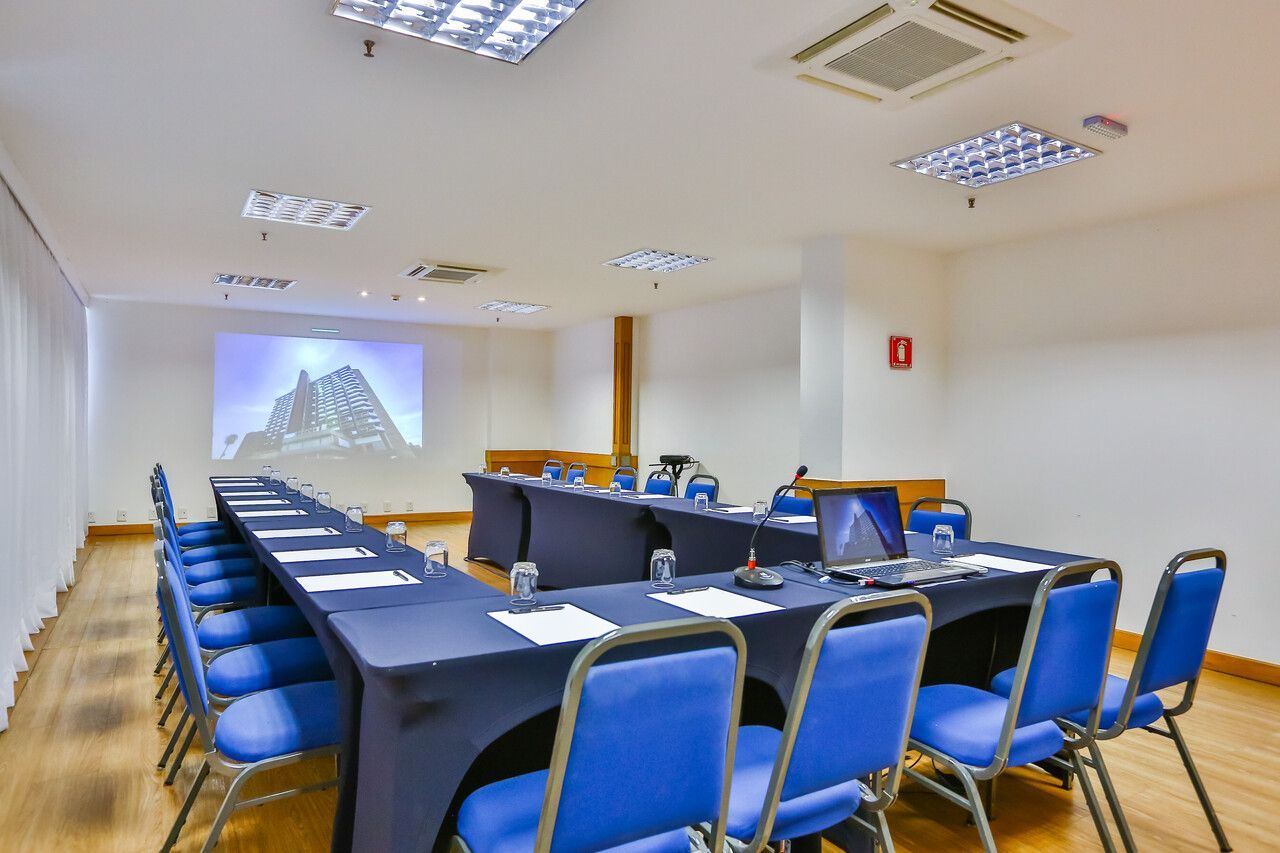 Sala de conferências com mesas azuis em formato de U, cadeiras, tela de projetor com imagem de montanha.