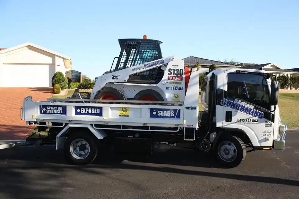A Dump Truck With A Bulldozer On The Back Is Parked In Front Of A House — Luke Seears Concreting in Port Macquarie, NSW