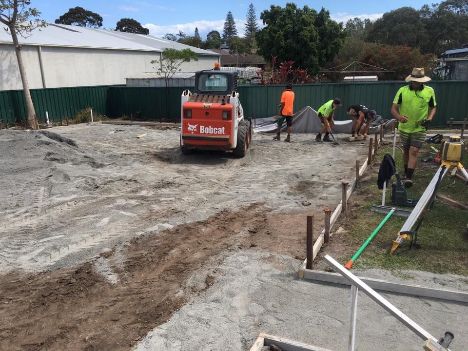 A Bobcat Is Driving Down A Dirt Road Next To A Group Of Construction Workers — Luke Seears Concreting in Laurieton, NSW