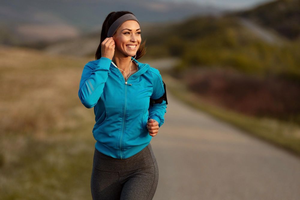 Woman jogging on a road in a blue jacket and black leggings, smiling in a scenic outdoor setting