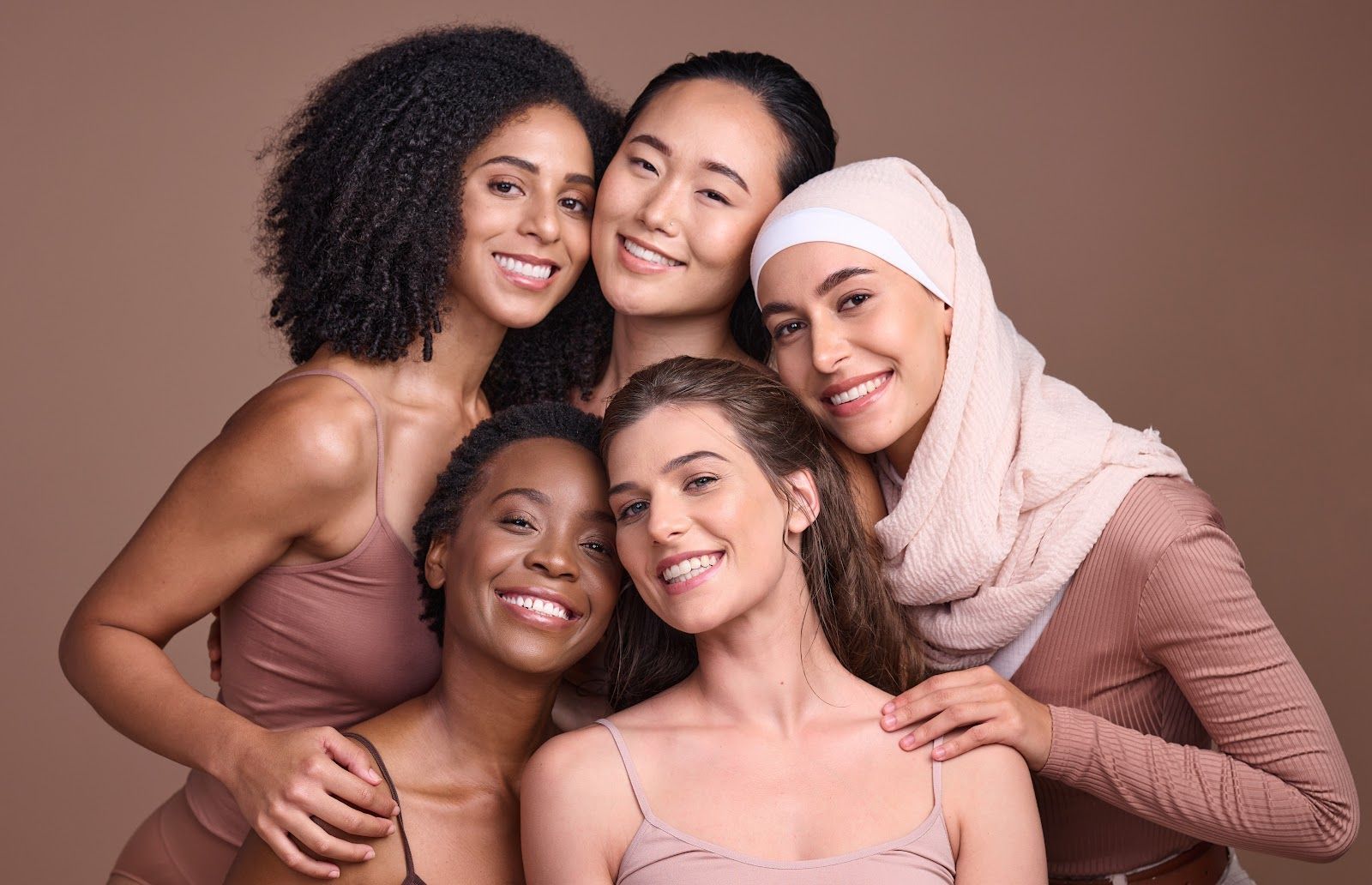 Five smiling women posing closely together against a soft pink background.