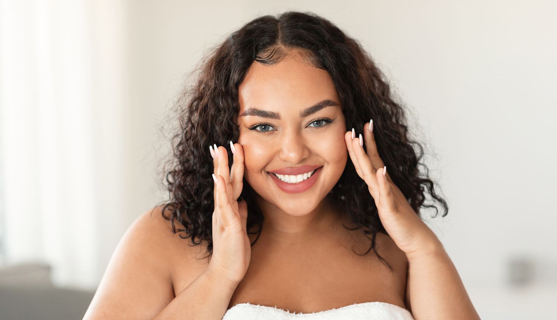 A woman with curly hair is smiling and touching her face with her hands.