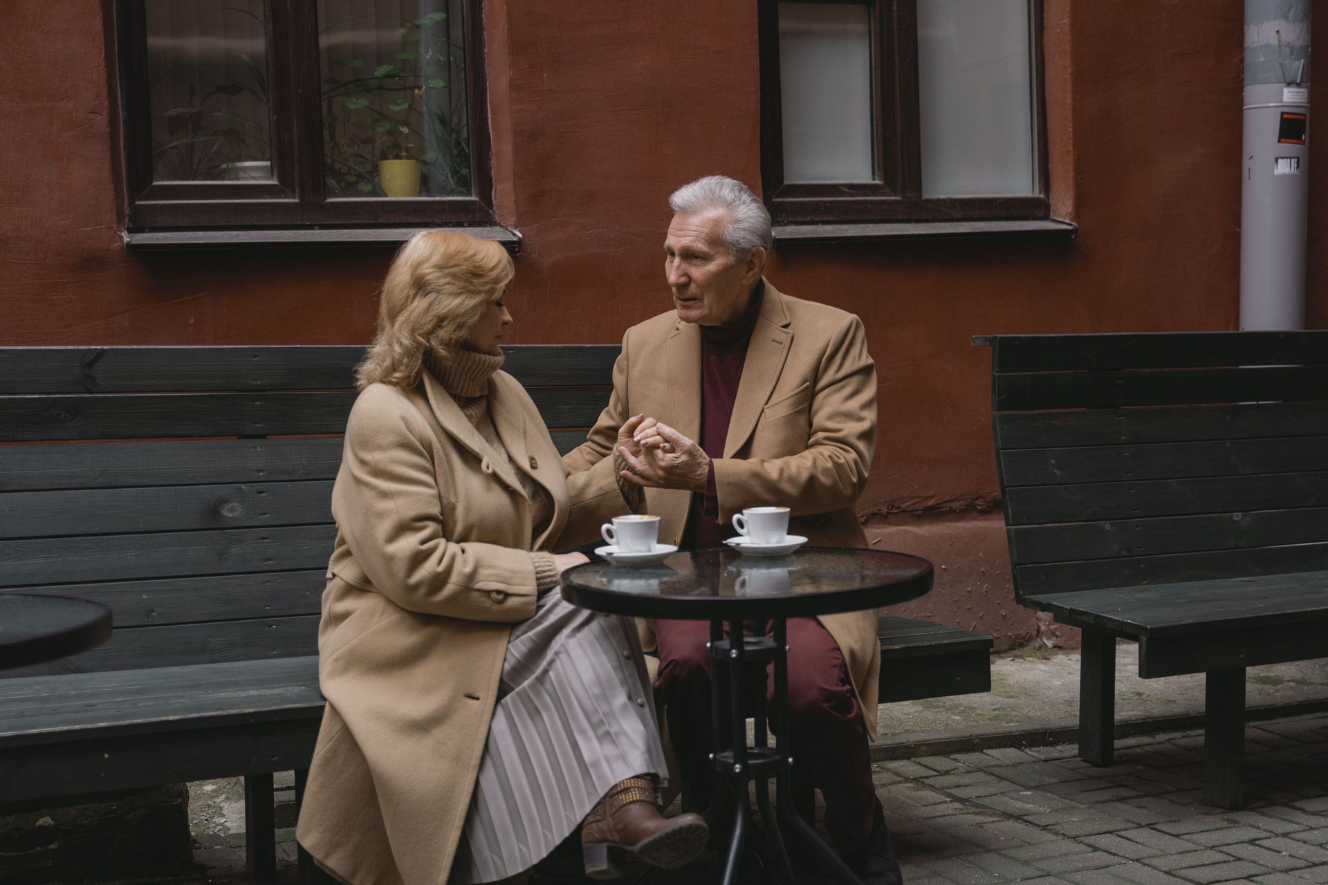 Two people chatting at a small outdoor café table, with coffee cups and a red brick wall behind them