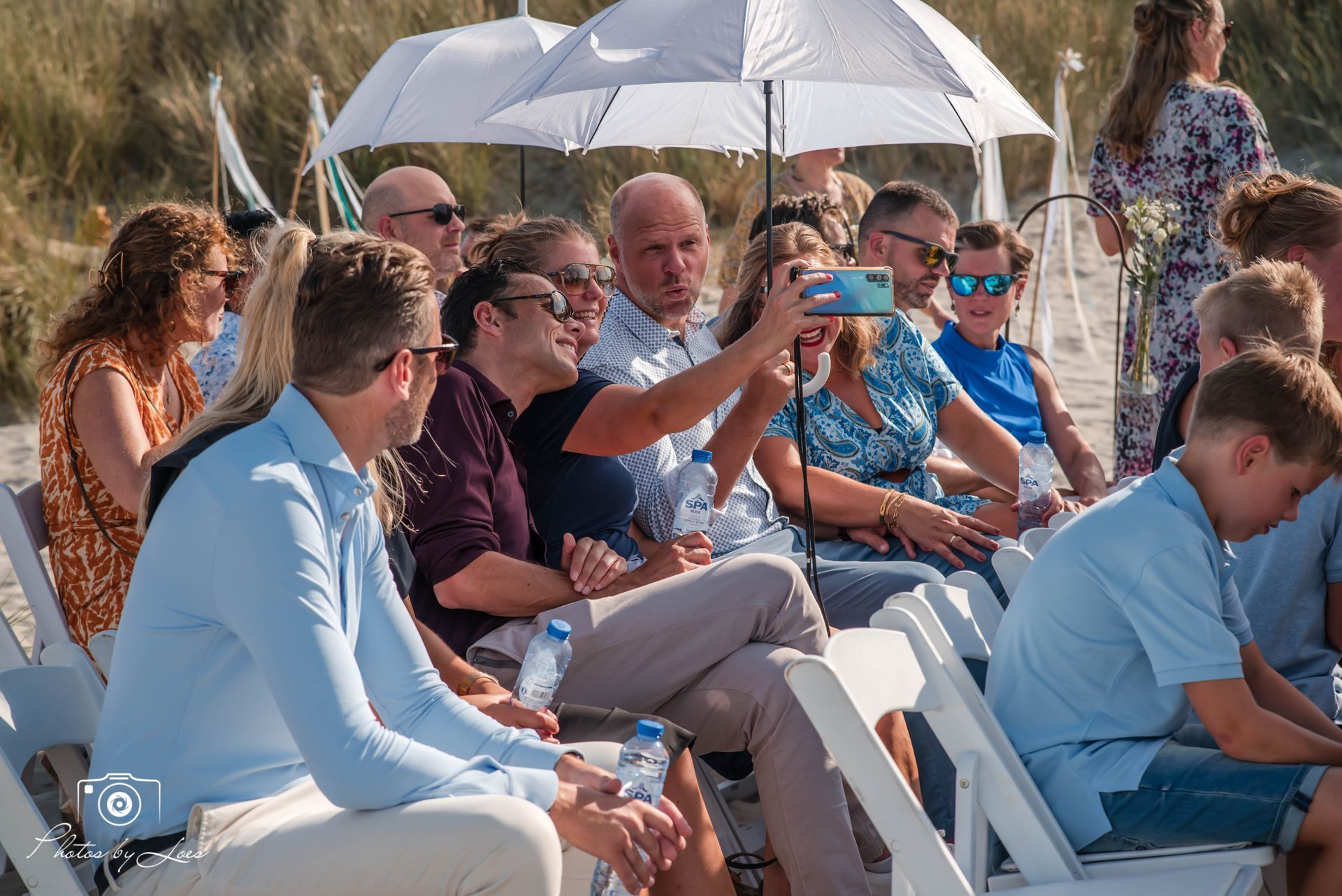 Een groep mensen zit op stoelen onder parasols en kijkt naar een huwelijksceremonie.