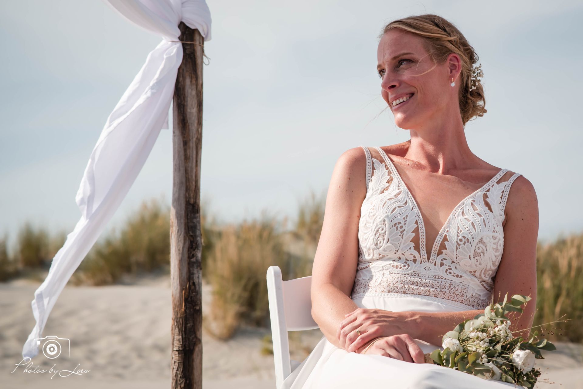 Een vrouw in een trouwjurk zit op een stoel op het strand.