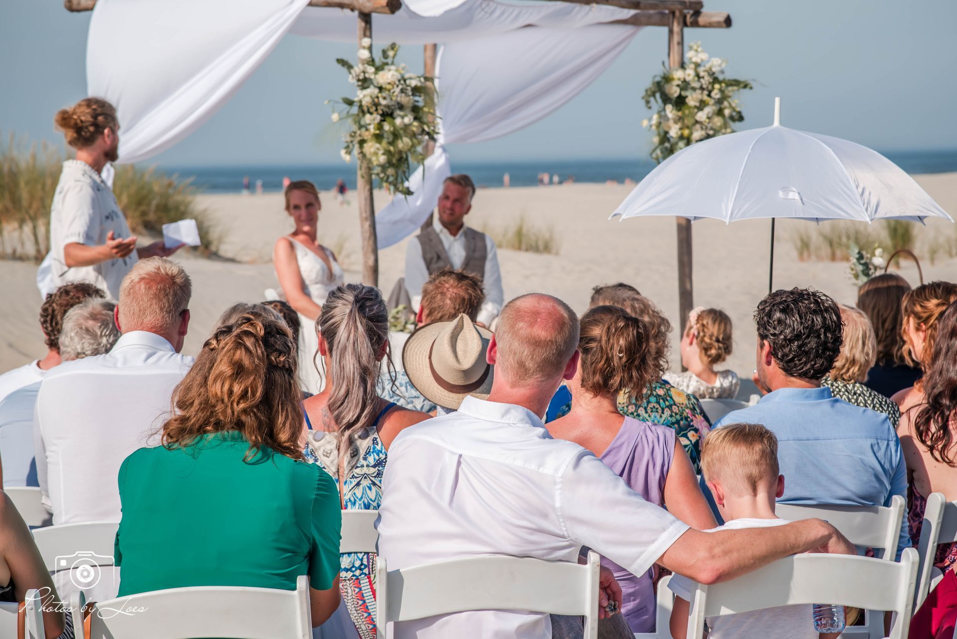 Een groep mensen zit op stoelen en kijkt naar een huwelijksceremonie op het strand.