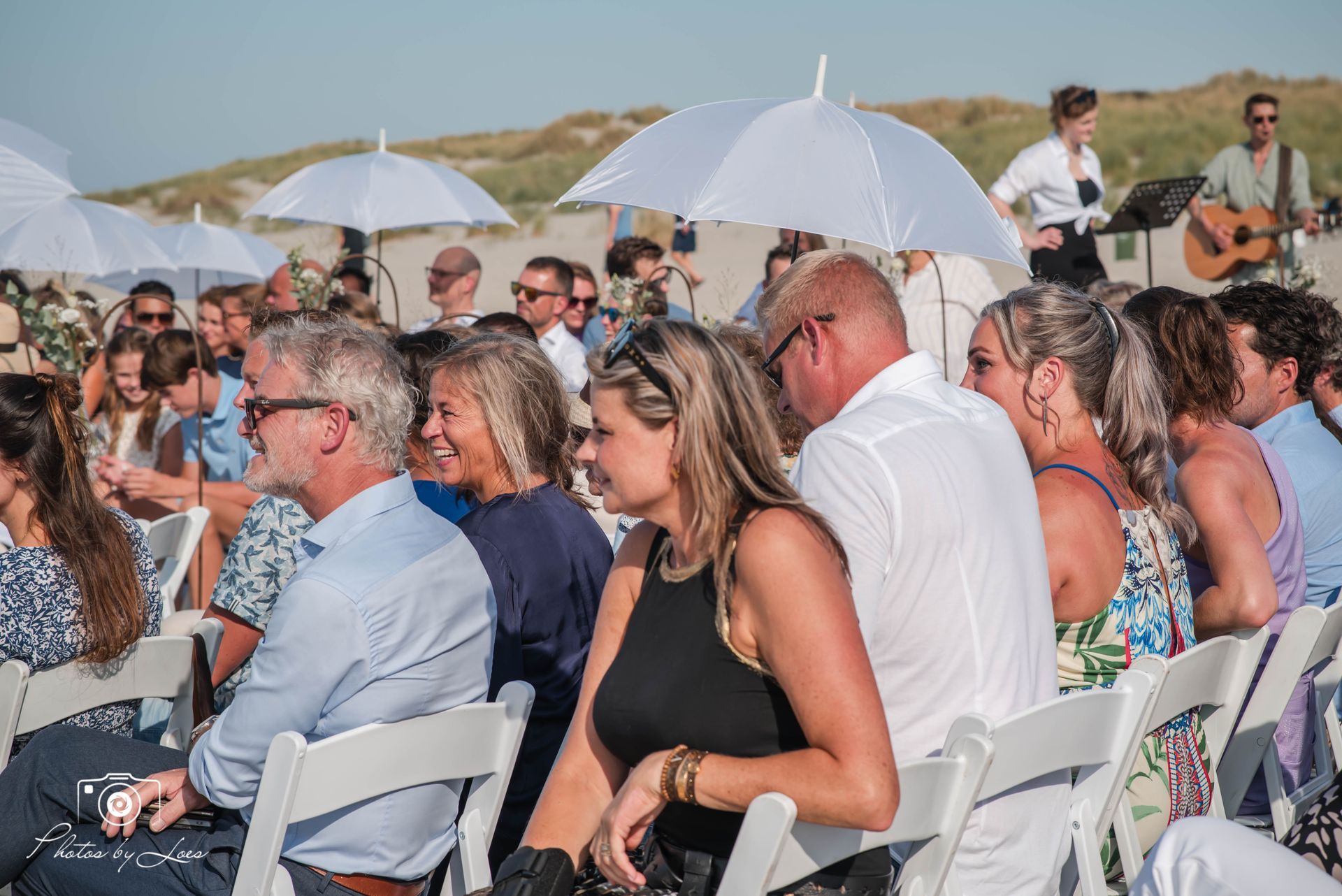 Een groep mensen zit op stoelen onder parasols tijdens een strandbruiloft.