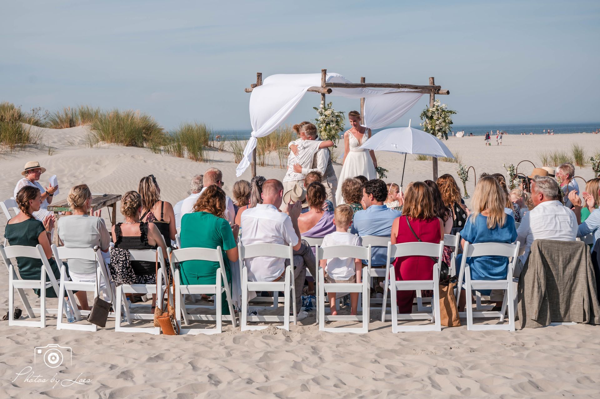 Een groep mensen zit op stoelen op het strand en kijkt naar een huwelijksceremonie.