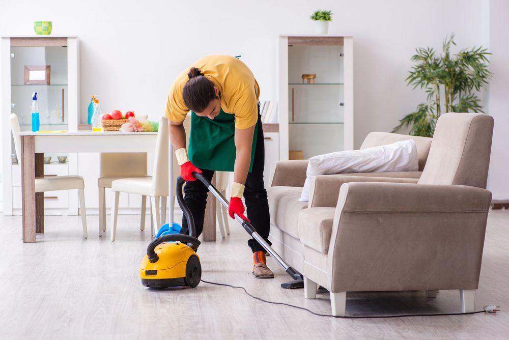 Una persona aspira una habitación con un sofá y una mesa. Lleva guantes y un delantal verde.