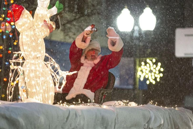Santa Claus waving from a decorated float beside a lit reindeer in falling snow