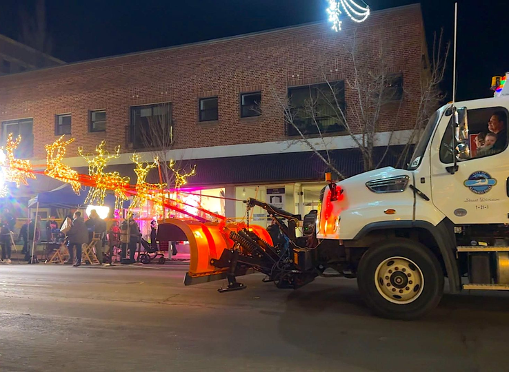 Night street parade with a fire truck, colorful lights, and a crowd beside a brick building