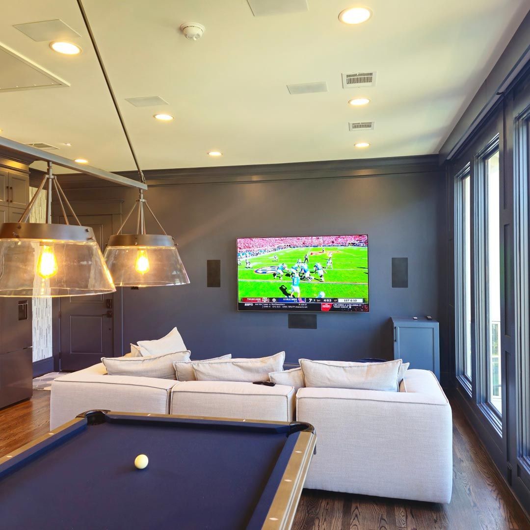 Modern living room with white sectional, pool table, wall TV, and large windows showing daylight