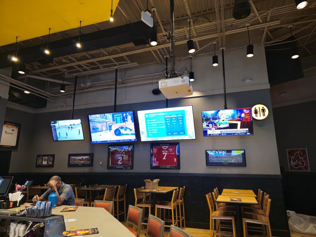 Sports bar interior with wall-mounted TVs, tables, stools, and a few patrons seated near the left.