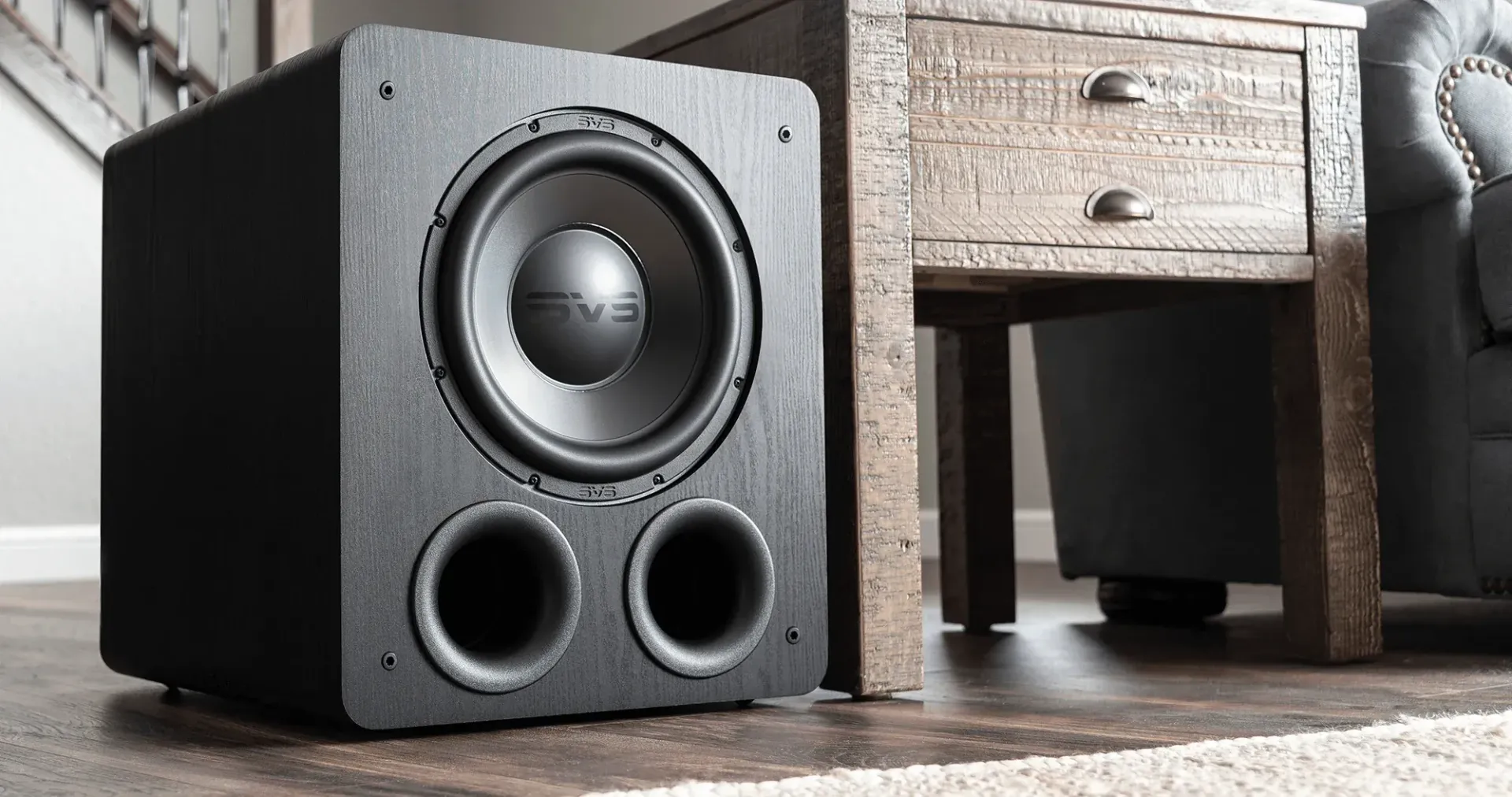 Black speaker on floor beside wooden cabinet, with large woofer and two bass ports.