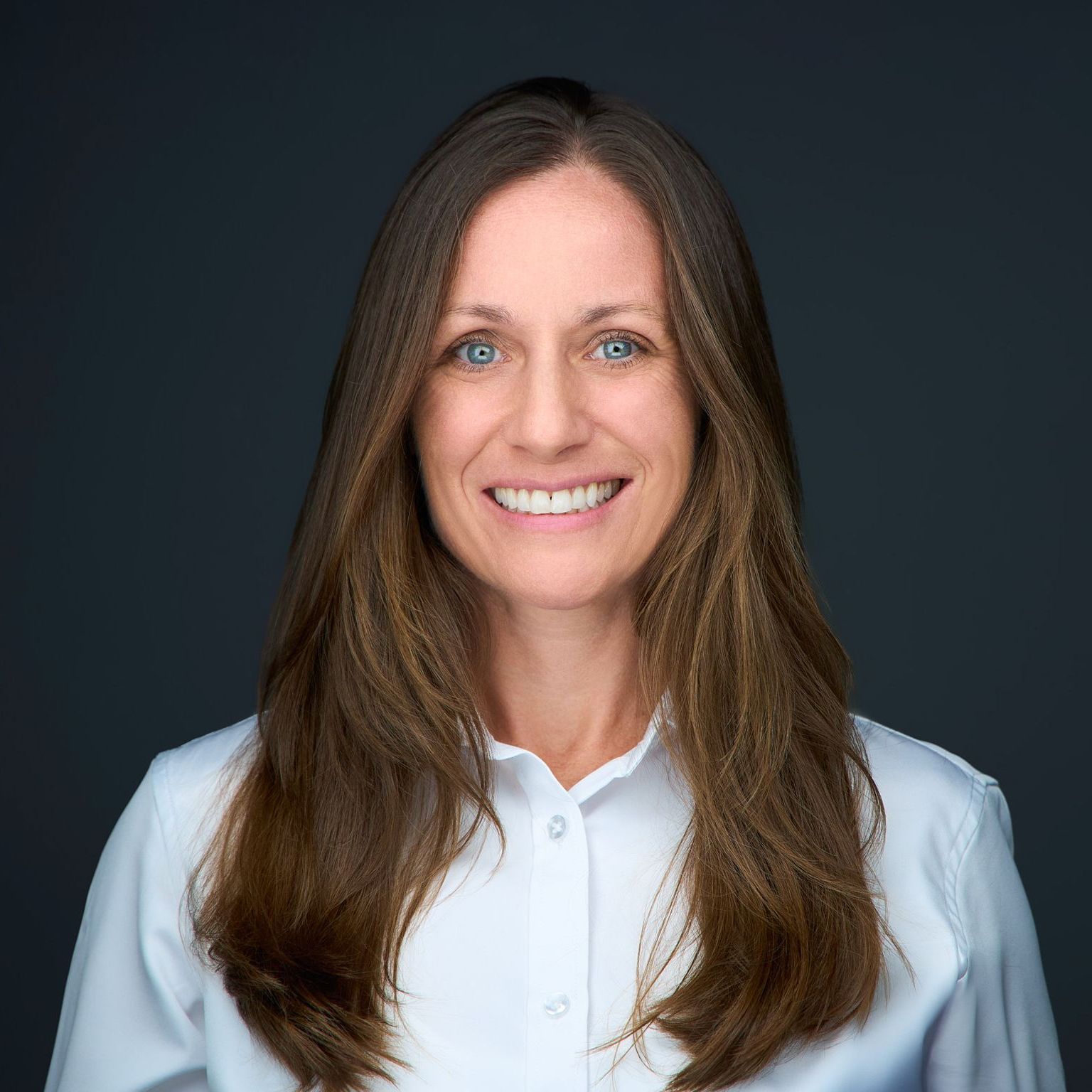 A woman with long brown hair is wearing a white shirt and smiling.