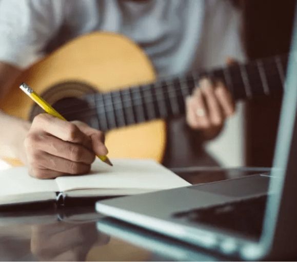 A person is writing in a notebook while playing a guitar.