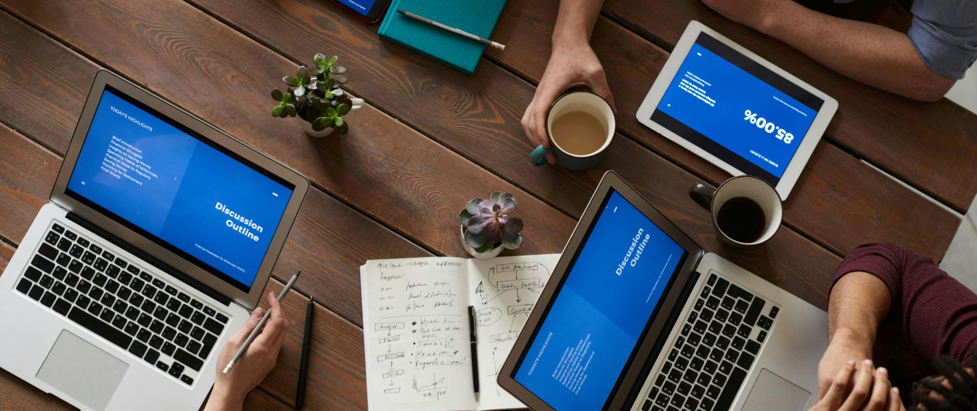 People working collaboratively at a wooden table, using laptops, tablets, and taking notes.