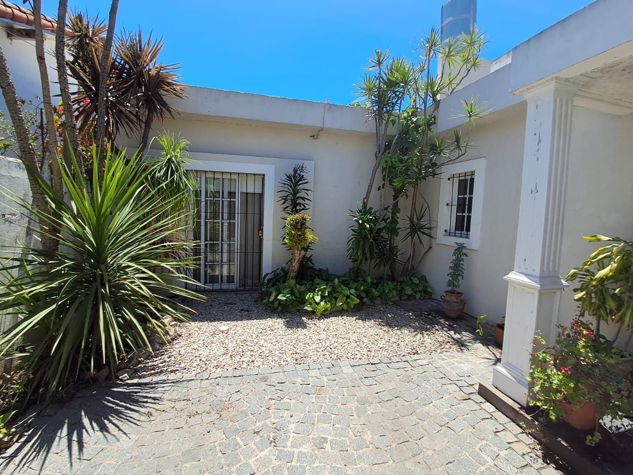 Patio blanco con macetas, sendero de grava y un cielo azul brillante junto a una casa de estuco.
