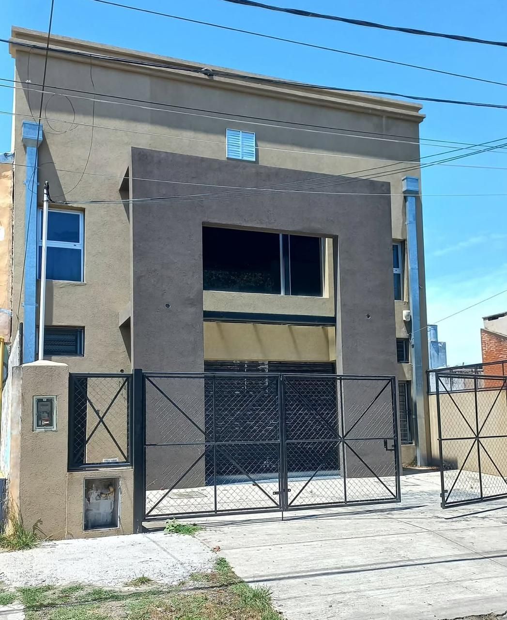 Casa beige de dos pisos con portón y cerca de metal negro bajo un cielo azul claro.
