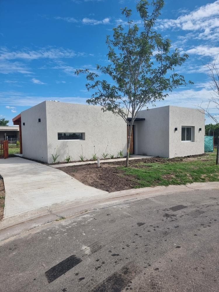 Pequeña casa moderna de color blanco con techo plano, entrada para vehículos y un árbol joven bajo un cielo azul.