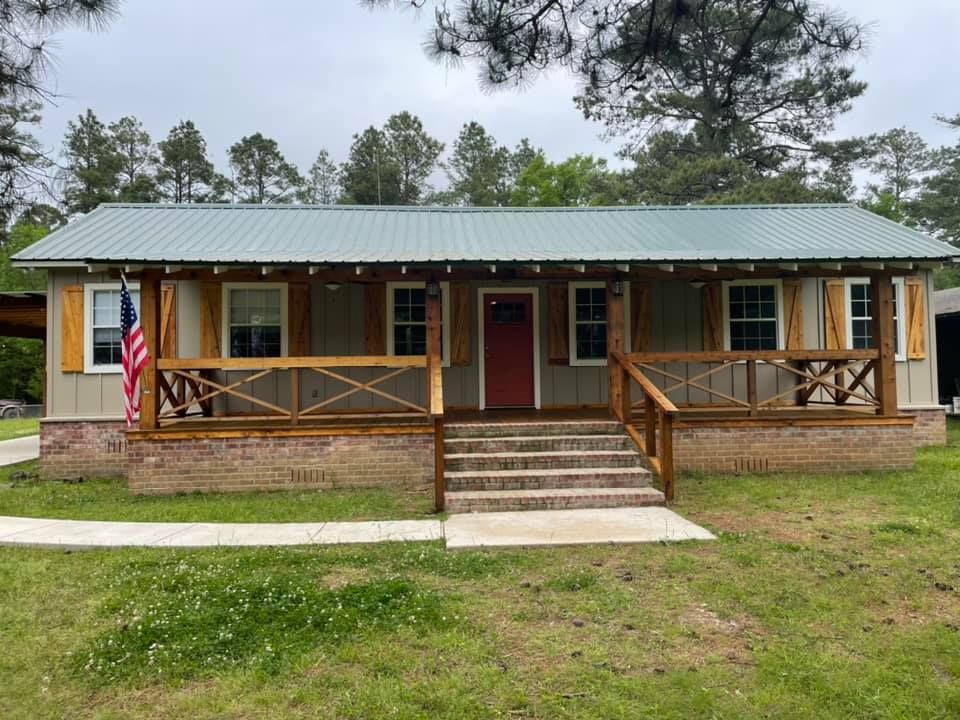 Single-story cabin with green roof, red door, and front porch, set in a grassy yard with an American flag.