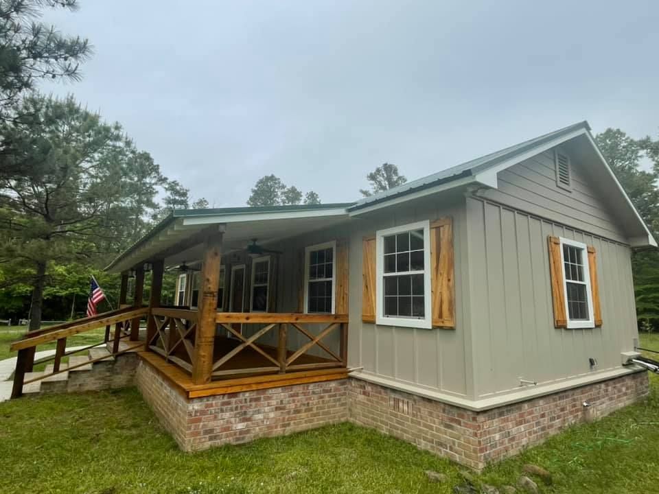 Small beige cabin with a wood porch and stone foundation in a wooded setting