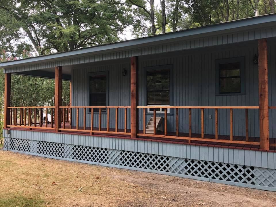 Covered porch on a gray cabin with wooden railings and lattice skirting, surrounded by trees.