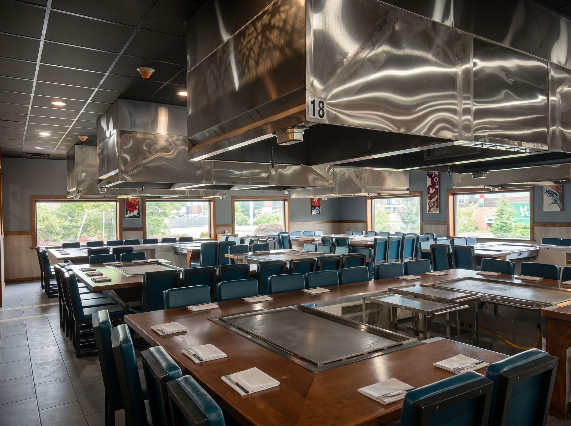 Modern restaurant dining room with blue chairs, wooden tables, and large windows under stainless steel vents