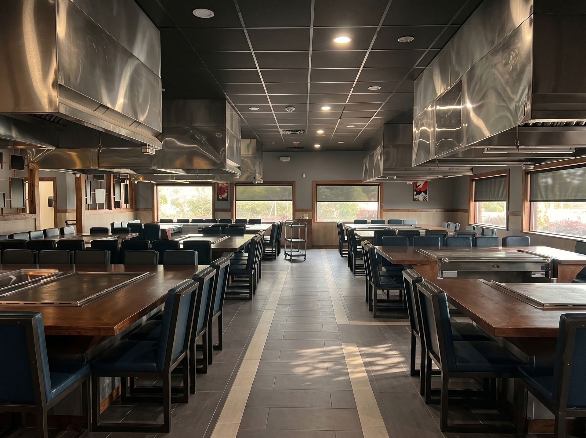 Empty restaurant dining room with long wooden tables, black chairs, and a dark ceiling.