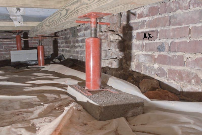 Crawlspace under a wooden floor with red support jacks, plastic sheeting, and a brick wall on the right