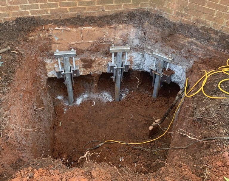 Excavation beside a brick wall with three metal support jacks and a yellow cord in a dirt pit