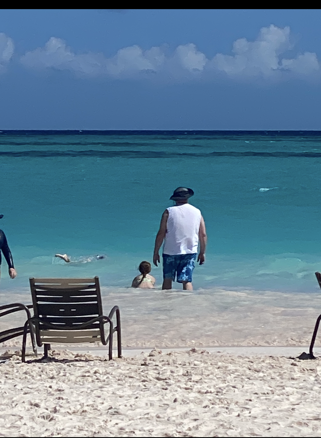 Beach scene with a chair on white sand, people at the turquoise shoreline, and a blue sky with clouds.