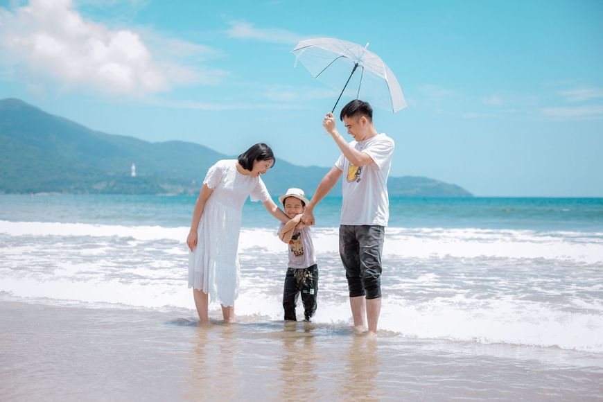 Family standing in shallow surf under a clear beach umbrella at a sunny seaside beach