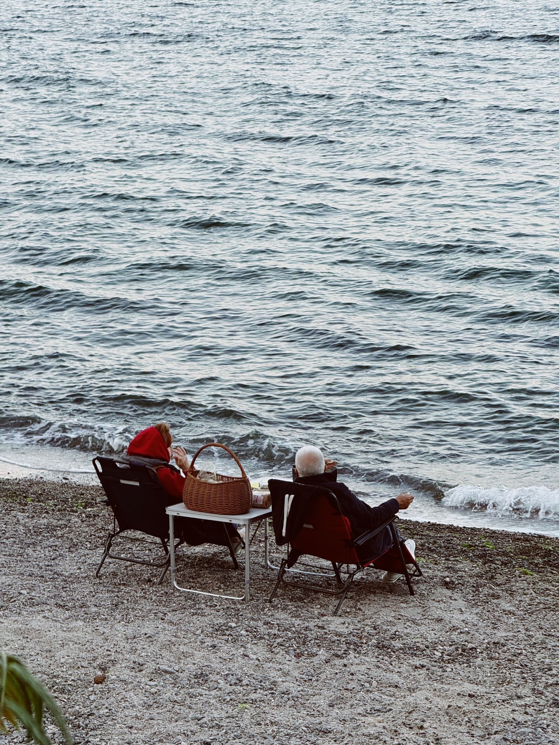 Two people in red chairs sit by a rocky shoreline, looking out at the calm sea.