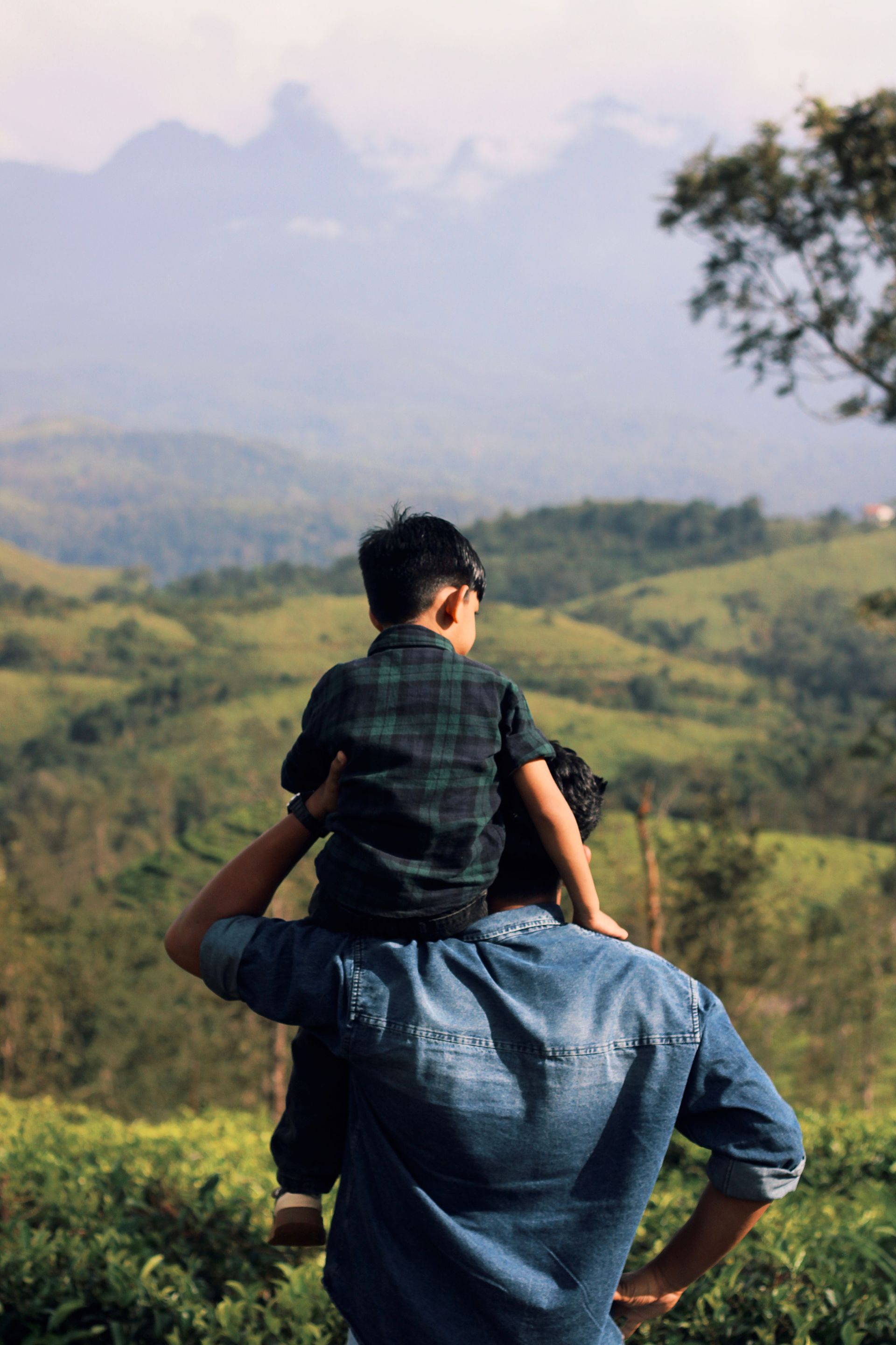 Adult carrying a child on shoulders, overlooking green hills and mountains.