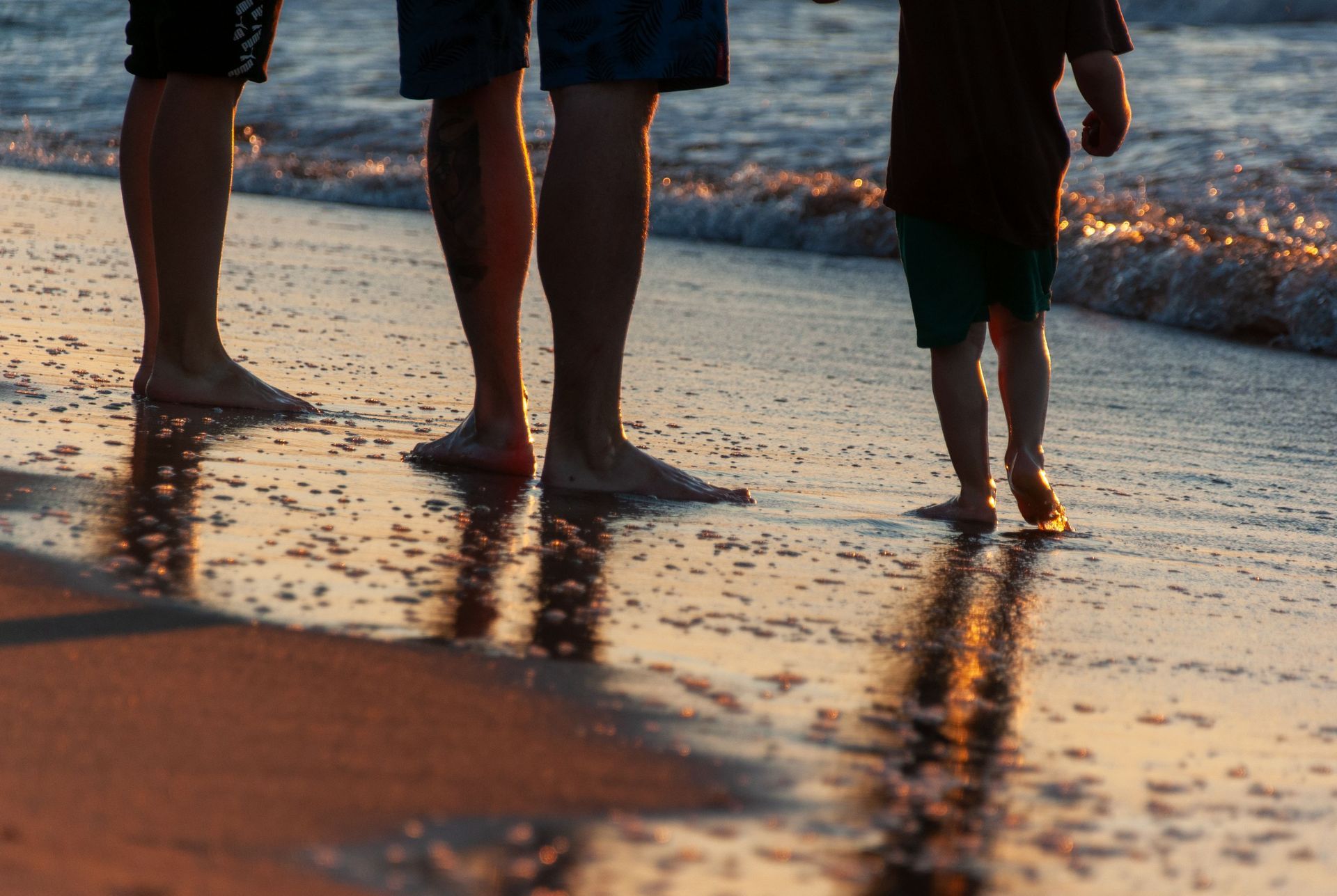 Three people walking barefoot along a wet beach at sunset, with waves and reflections on the sand