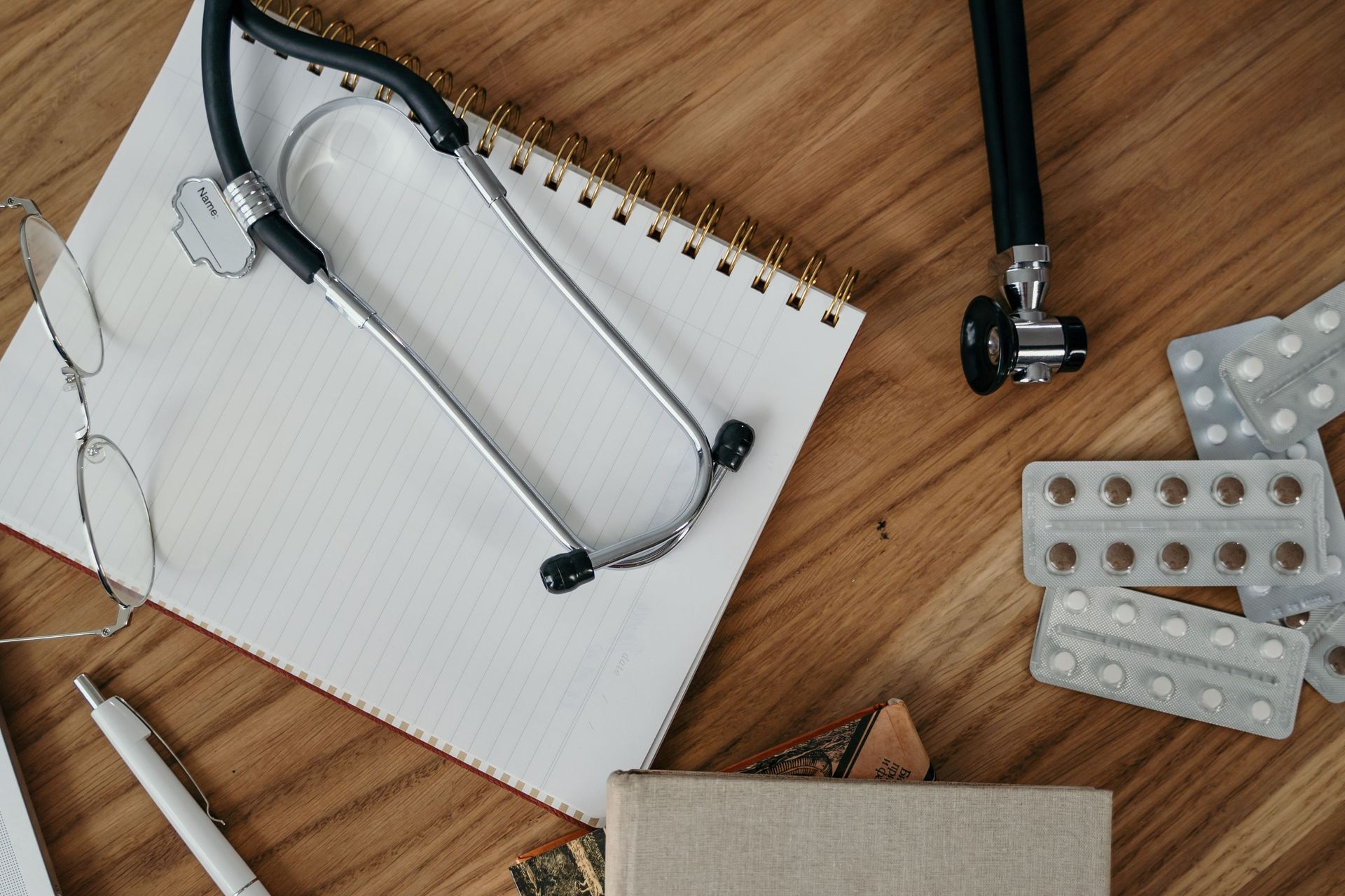Notebook with stethoscope, medicine blister packs, and eyeglasses on a wooden desk