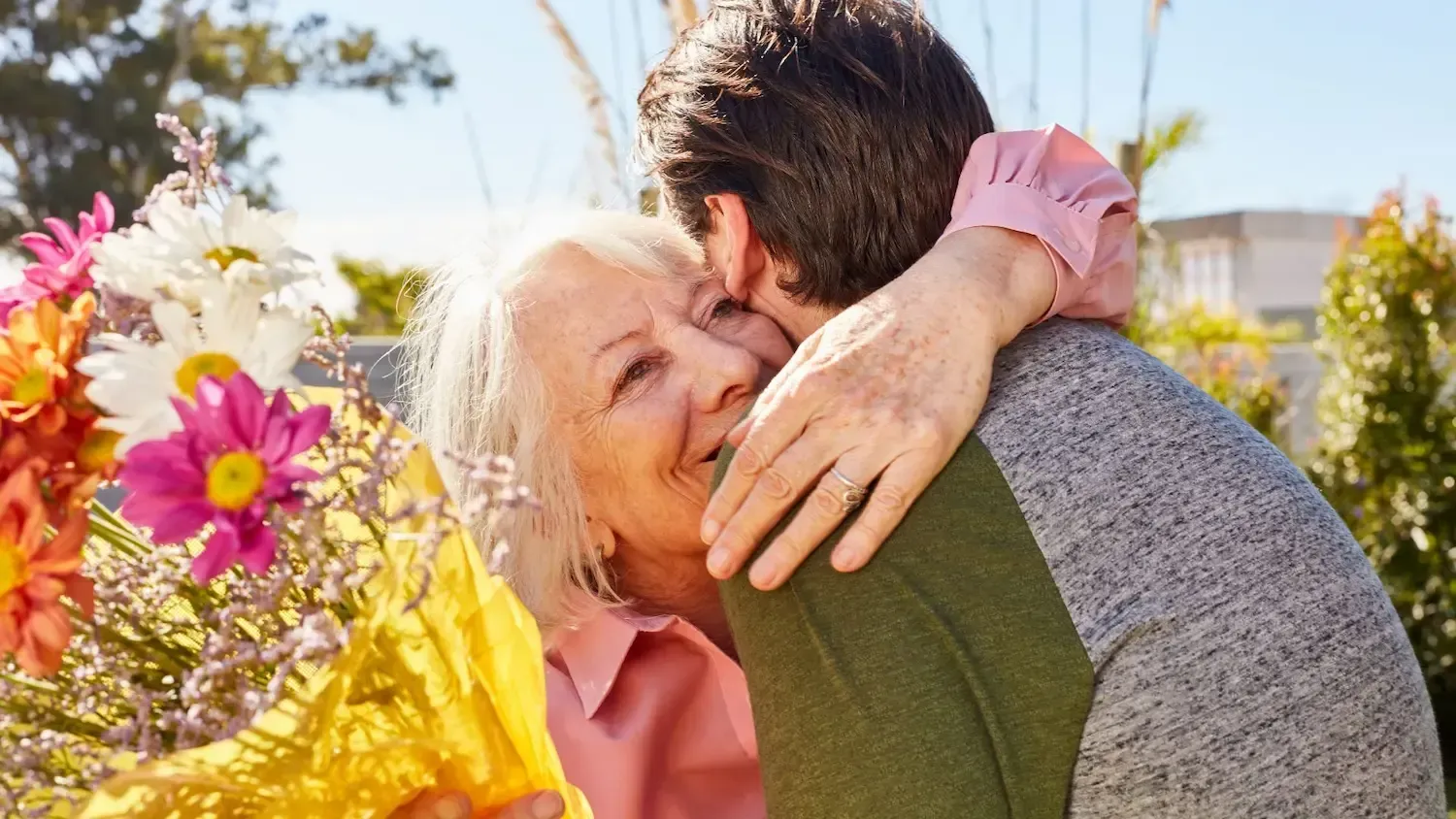 Woman hugs a person outdoors, holding flowers. She smiles, embracing. Sunny day.