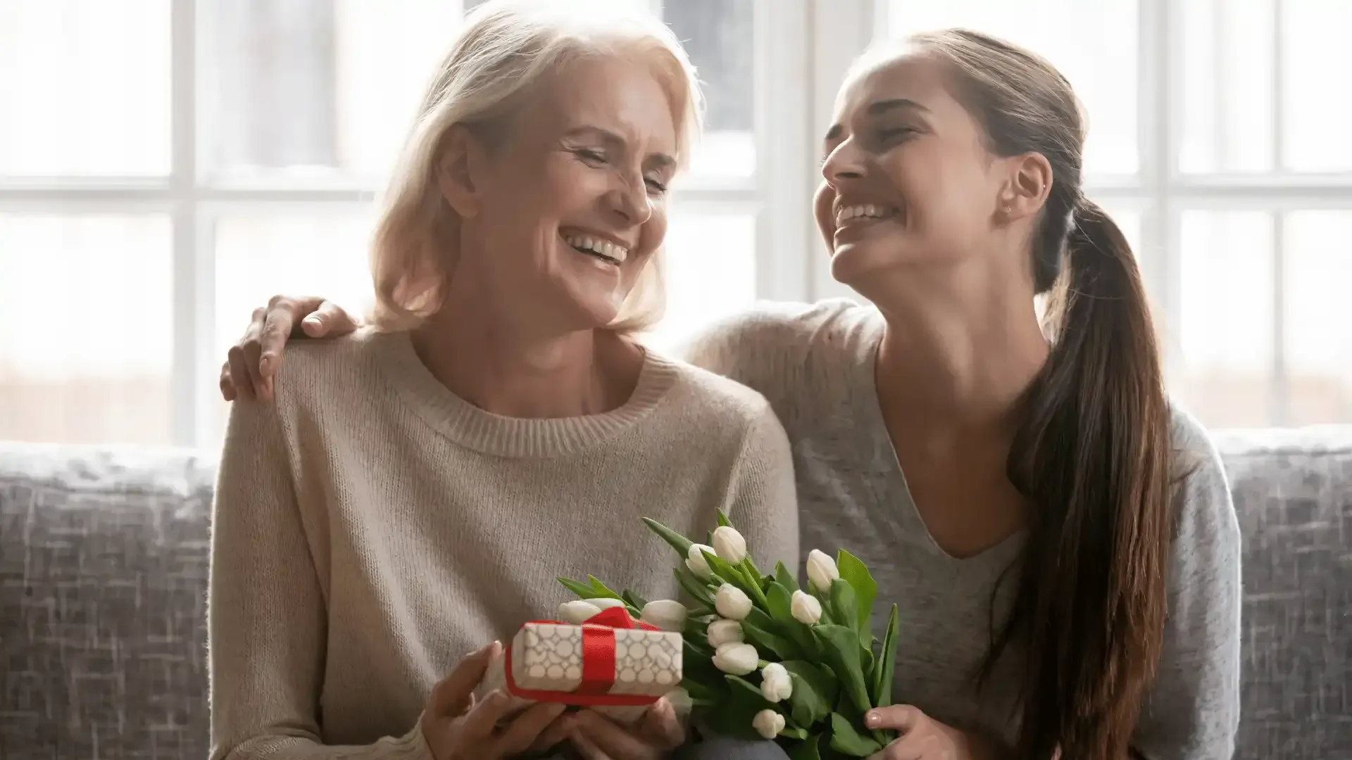 Two women are sitting on a couch holding flowers and a gift.