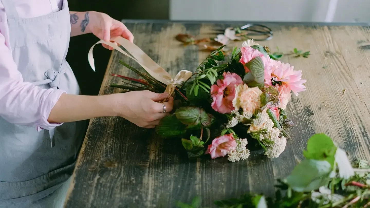 Person tying a ribbon around a bouquet of pink and white flowers on a wooden table.