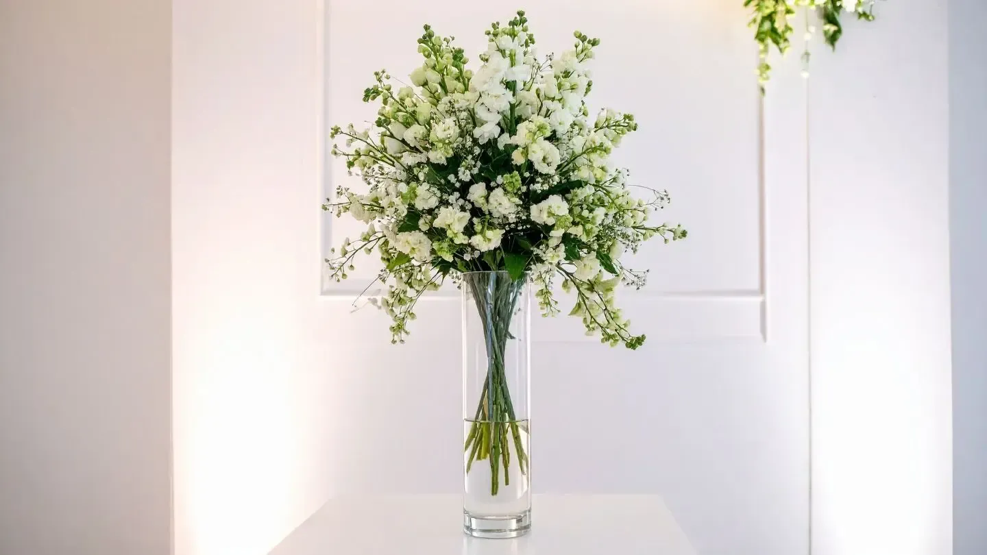 White flowers in a tall glass vase on a white table, set against a white wall.