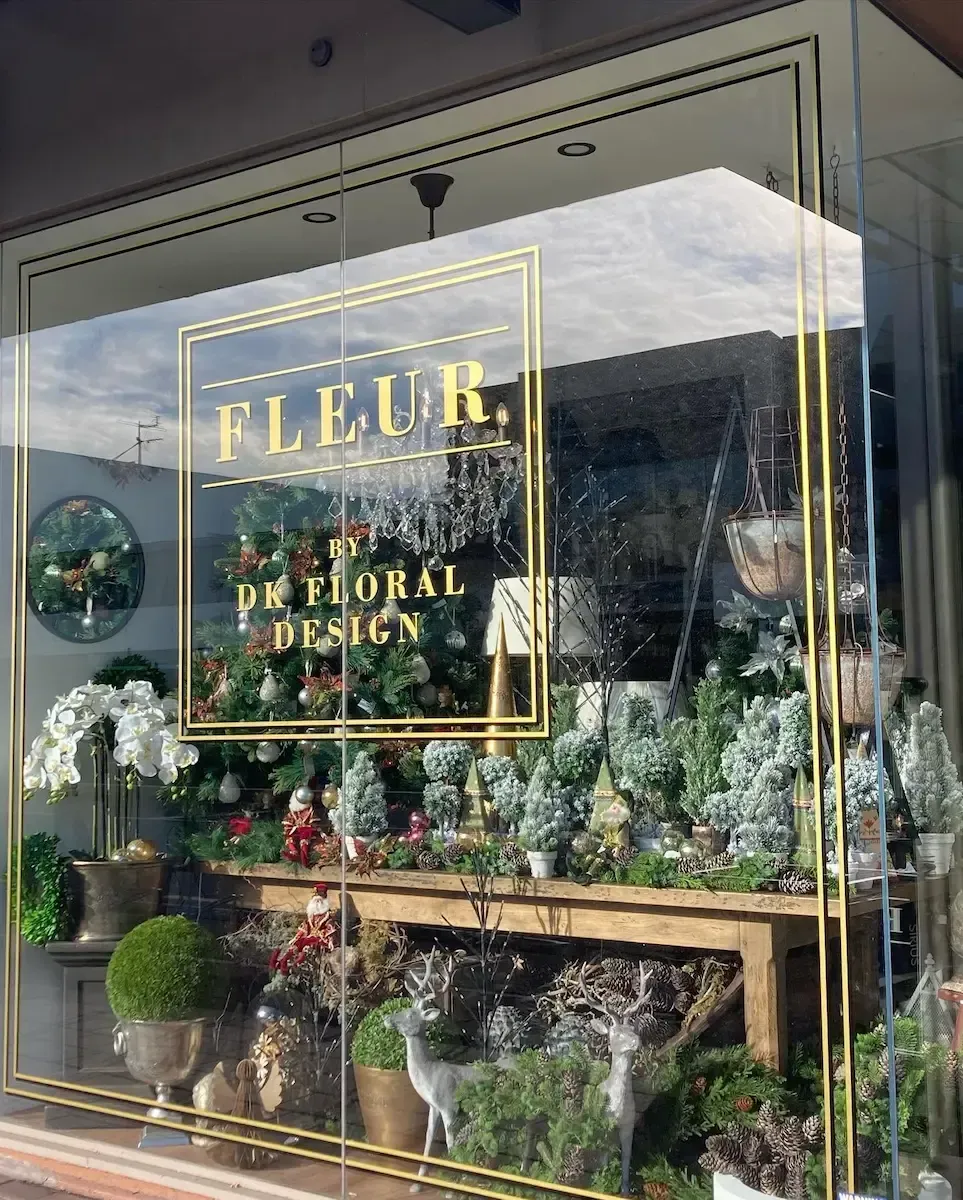 Flower shop window display with gold “FLEUR” lettering, floral arrangements, and potted plants.