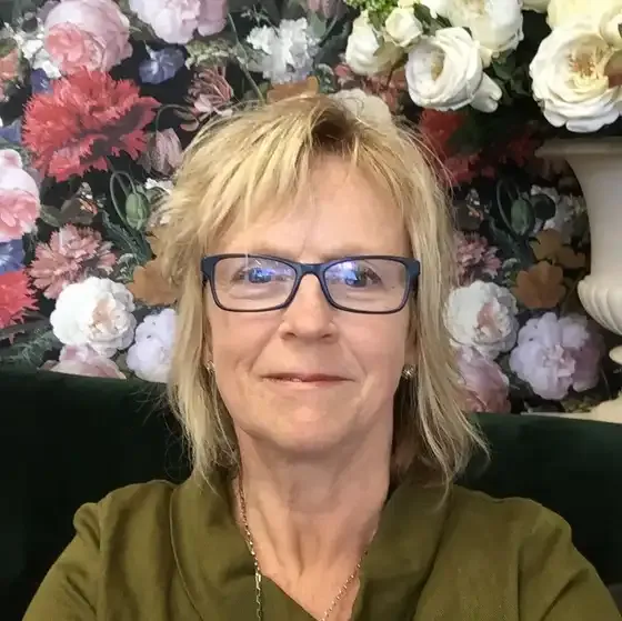Debra wearing glasses is sitting in front of a floral wallpaper .