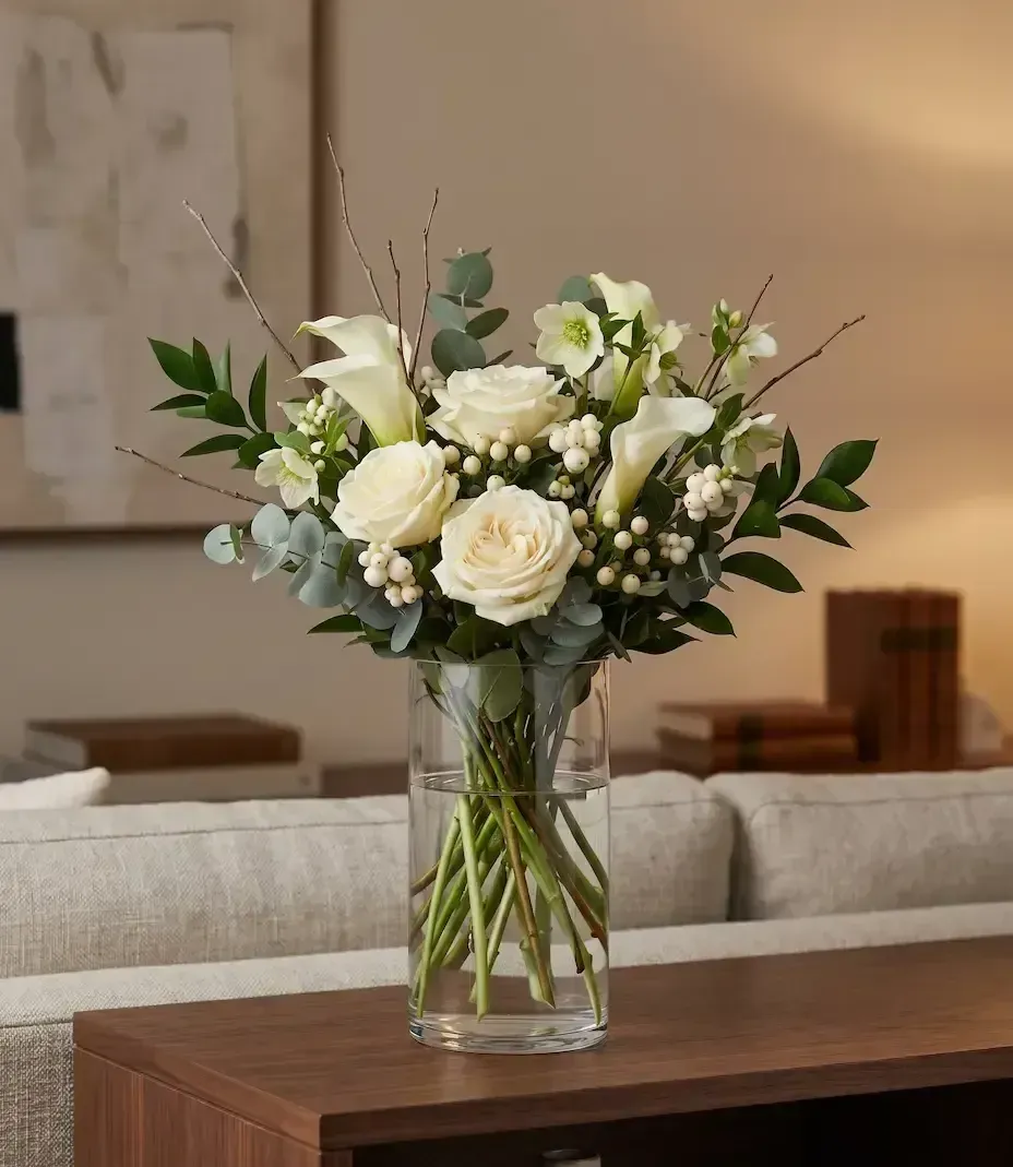 White roses and greenery in a glass vase on a wooden table in a living room