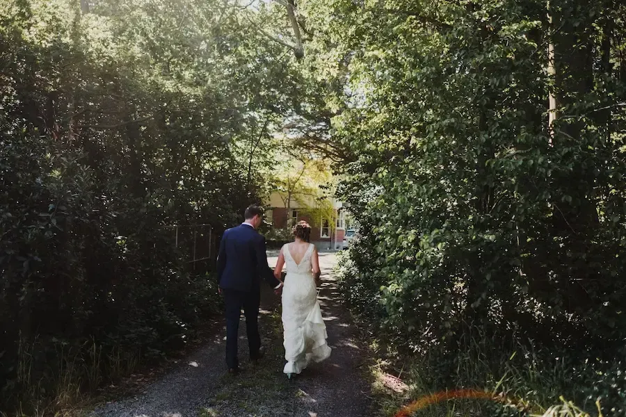 Couple walking on path lined by trees toward a building. Sunlight streams through the foliage.