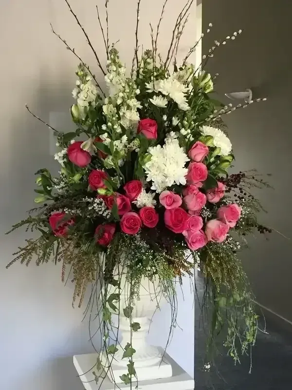 Large floral arrangement in a white urn featuring pink roses, white snapdragons, hydrangea, and willow branches.