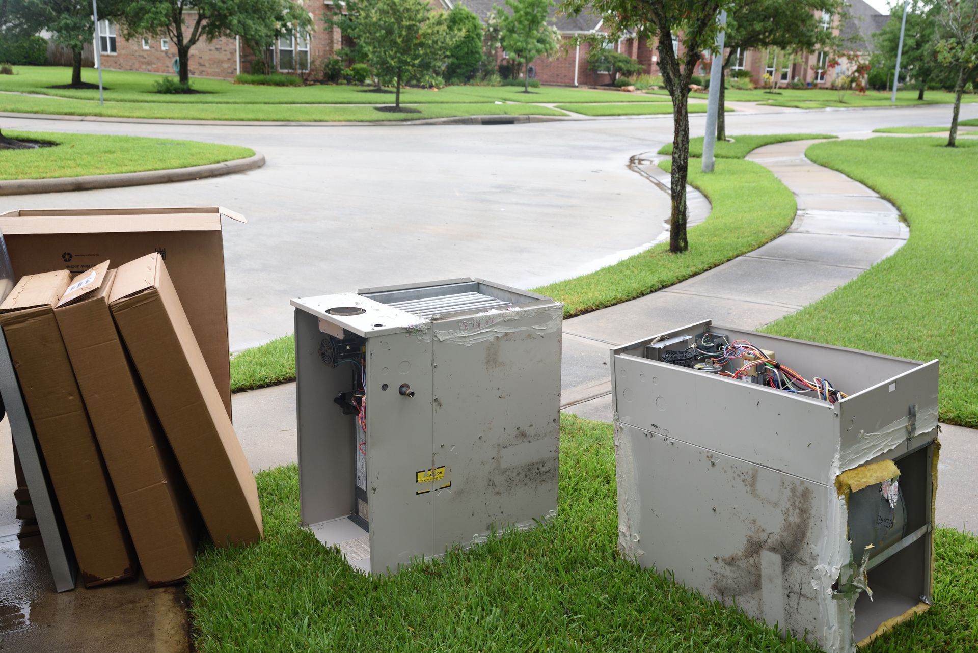 Old air conditioning units and cardboard boxes on a lawn near a sidewalk and houses.