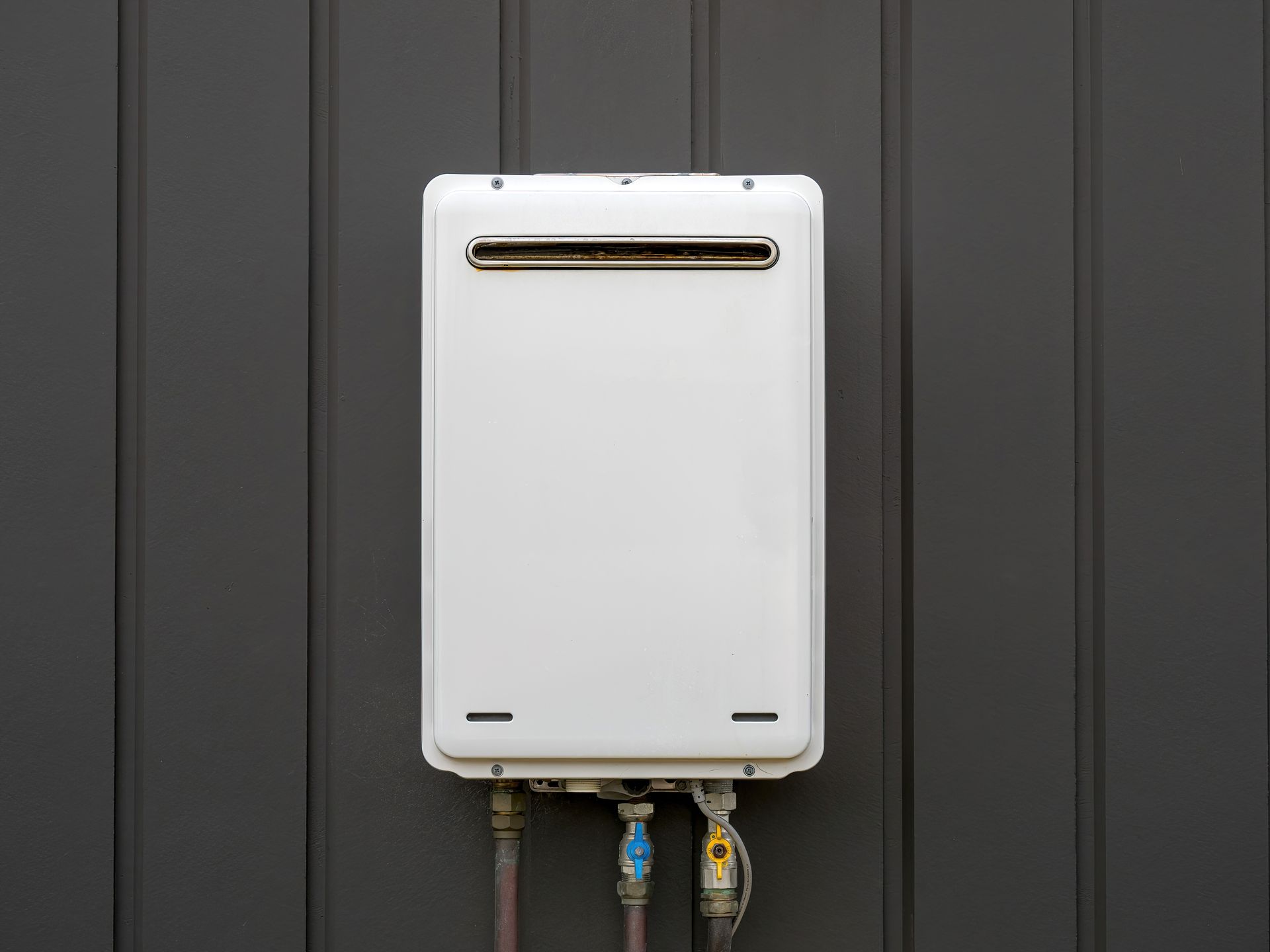 White tankless water heater mounted on a dark gray exterior wall with pipes.